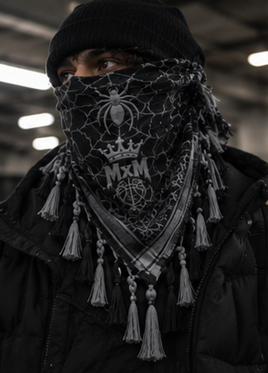 MxM black and grey spiderweb keffiyeh on a model with prominent spider face motif, crowned MxM basketball logo and thin knotted tassels. Digital design preview, not a manufactured sample photo.
