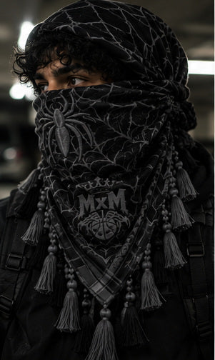 MxM black grey keffiyeh on a model with spider face, web pattern, crowned MxM emblem and hanging tassels. Digital preorder design mockup, not a physical sample photo.