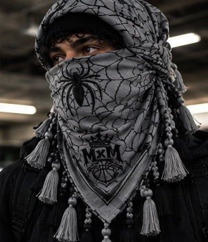 MxM silver grey keffiyeh on a model with spider face, web pattern, crowned MxM emblem and hanging tassels. Digital preorder design mockup, not a physical sample photo.