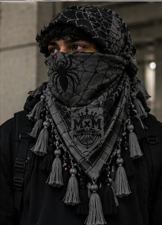 MxM webbed keffiyeh charcoal black model front. Crowned MxM logo appears on front only; back features woven web pattern and knotted fringe; photorealistic preorder design mockup.