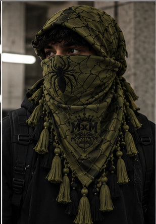 MxM webbed keffiyeh olive black model front. Crowned MxM logo appears on front only; back features woven web pattern and knotted fringe; photorealistic preorder design mockup.