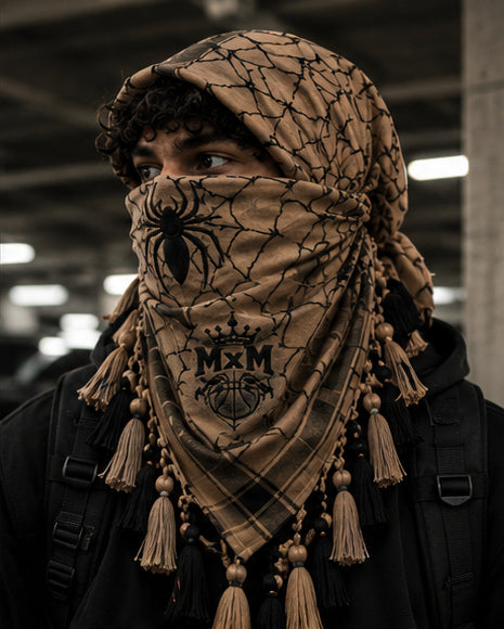 MxM webbed keffiyeh tan black model front. Crowned MxM logo appears on front only; back features woven web pattern and knotted fringe; photorealistic preorder design mockup.